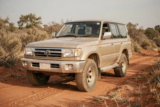 Toyota Land Cruiser 100 HDJ d'occasion garé sur une piste de terre rouge entouré de végétation sèche, vue grand-angle trois-quarts avant